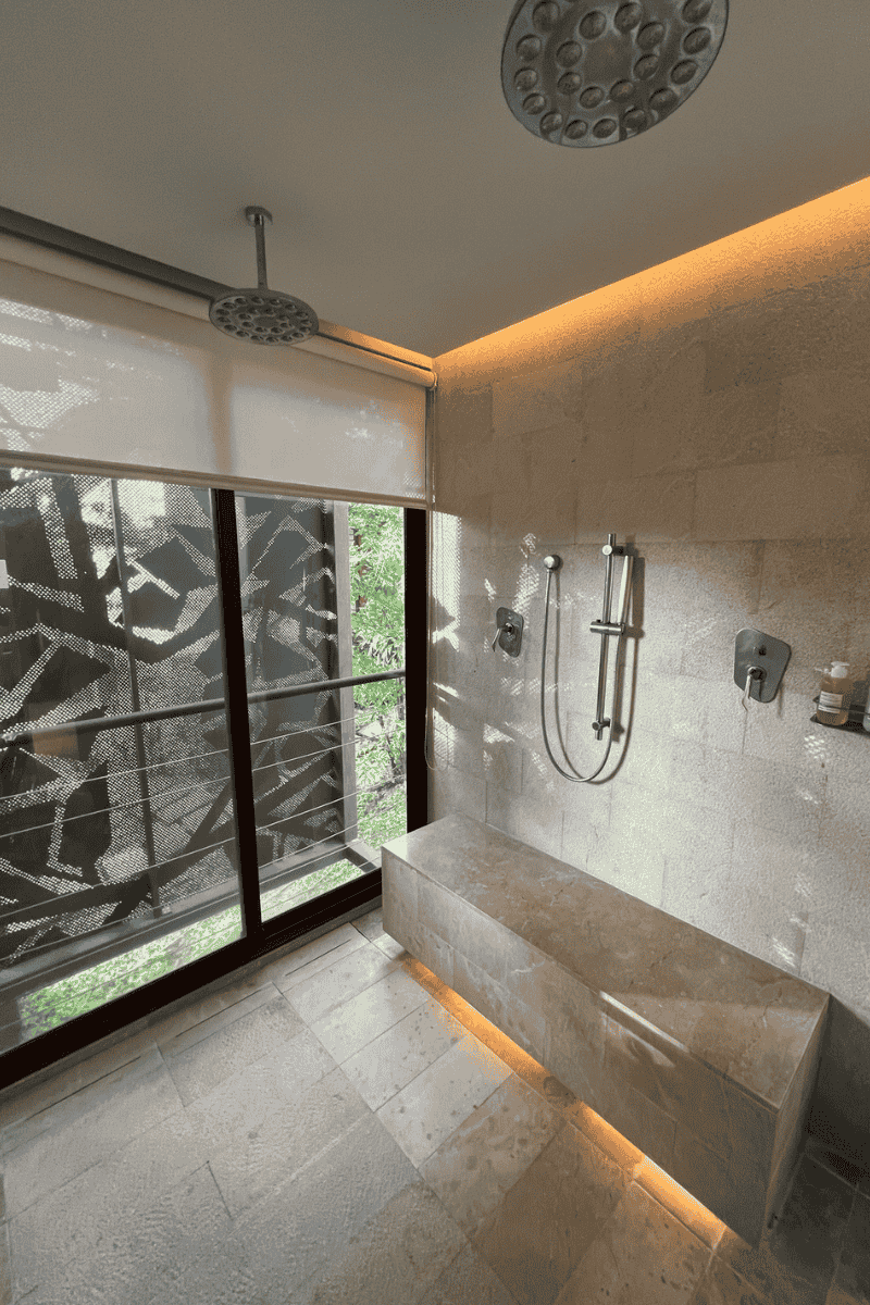 Modern bathroom with large shower, dual showerheads, and a stone bench. Floor-to-ceiling window with patterned blinds partially open, bathing the room in natural light.